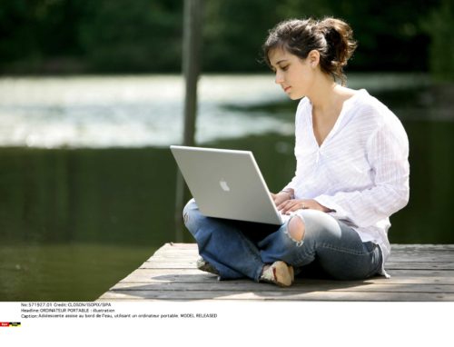 Adolescente assise au bord de l'eau, utilisant un ordinateur portable pour consulter ses newsletters